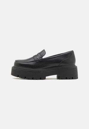 Mocassins en cuir noir avec une semelle en caoutchouc épaisse et basse, des détails cousus sur le dessus et une texture lisse.