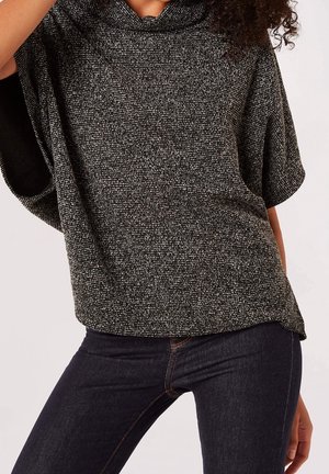 Vrouw met een losse grijze gestructureerde korte mouwen trui met een col en donkere slim-fit jeans tegen een effen achtergrond.