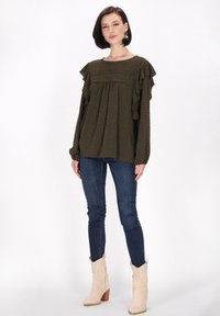 Oliwkowa bluzka z falbanami na rękawach, teksturowana tkanina, okrągły dekolt i mały wzór w kropki, w połączeniu z ciemnymi dżinsami i beżowymi botkami na obcasie.
