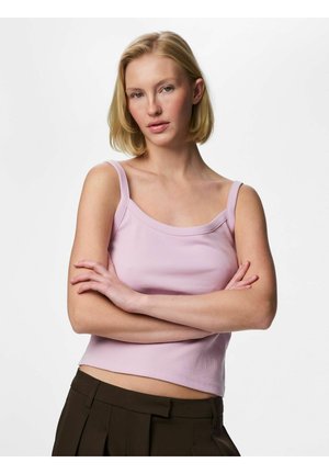 Femme blonde aux cheveux courts portant un débardeur violet clair et un pantalon foncé, les bras croisés, sur fond blanc.