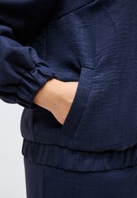 Χέρι τοποθετημένο στην τσέπη ενός σκούρου μπλε σακακιού με ελαστικό μανσέτα και ζώνη μέσης, που δείχνει την υφή του υφάσματος και τις λεπτομέρειες της ραφής.