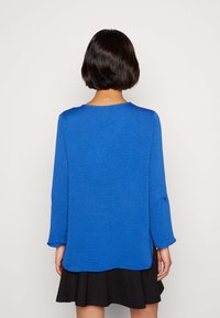 Niebieska bluzka z długim rękawem o teksturowanej tkaninie, z okrągłym dekoltem i bocznymi rozcięciami. W połączeniu z czarną spódnicą.