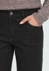 Jeans de mezclilla negra con un bolsillo frontal, cierre de botón de latón y una textura sutil. Combinados con un suéter ligero y texturizado.