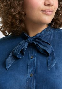 Jeansowa koszula w głębokim niebieskim kolorze, z zawiązywanym kołnierzykiem, guzikiem z przodu oraz lekko teksturowanym materiałem. Posiada małe brązowe guziki.