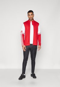 Rode sportjack met witte mouwen, ritssluiting en drie strepen op de schouders; gecombineerd met zwarte, aansluitende broek en zwarte schoenen.