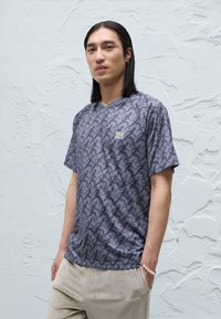 Šedé tričko so vzorom a jemným motívom vtáka, výstrih do V, krátke rukávy a malý logotyp pri hrudi.