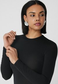 Černý body s dlouhým rukávem a přiléhavým střihem, s kulatým výstřihem, hladkým povrchem a jemným materiálem. Velké bílé výrazné náušnice.