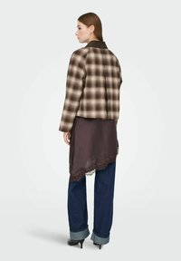 Kvinne med rett brunt hår iført brun rutete jakke over en asymmetrisk mørk brun kjole med blonder, oppbrettede blå jeans og svarte høyhælte sko, stående sideveis.