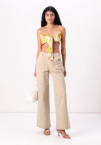 Top corto floreale con fiori gialli e arancioni, abbinato a pantaloni beige a gamba larga. La modella tiene una borsa bianca e indossa sandali con cinturino.