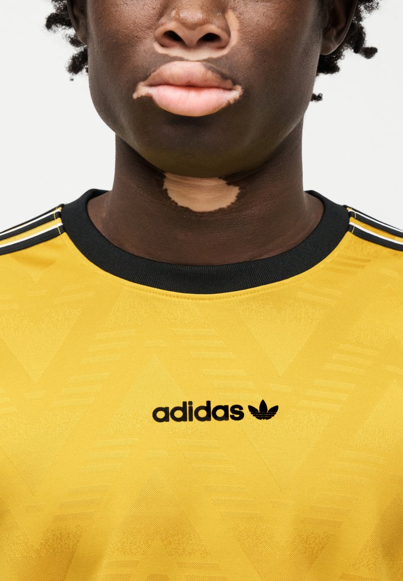 T-shirt de sport jaune Adidas avec un motif texturé, col noir et logo. Le design comprend des bandes noires sur les épaules.