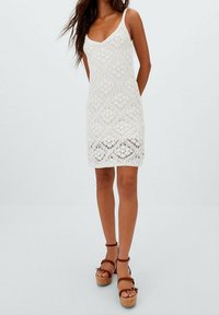 Robe blanche en crochet avec un motif géométrique, bretelles fines et silhouette ajustée. Associée à des sandales compensées marron.
