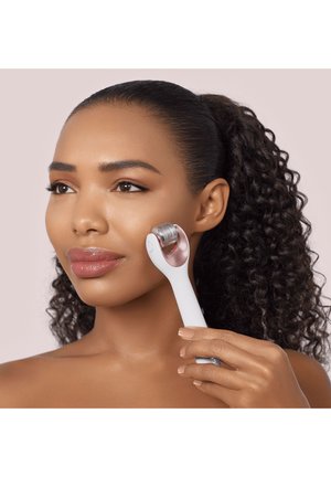 Woman with curly hair uses a white facial roller with metal needles on her cheek for skincare treatment.
