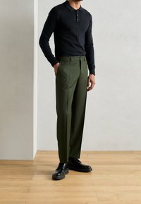 Polo a maniche lunghe nera con bottoni abbinata a pantaloni tailored verde oliva e scarpe stringate nere. Sfondo in legno.