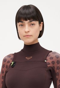 Costum de neopren lung, maro, cu manșete lungi și guler înalt, mâneci transparente cu model floral și un logo în formă de inimă pe piept. Accentuate cu detalii din metal.
