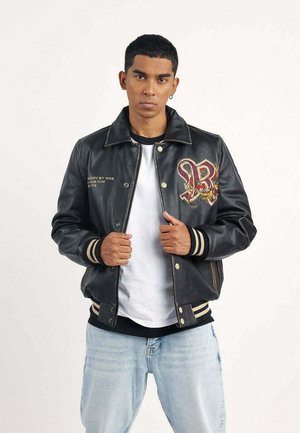 Jeune homme aux cheveux courts portant une veste en cuir noire avec un patch brodé "RB" et un jean bleu clair, tenant le col de la veste avec les deux mains.