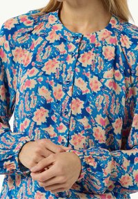 Blusa de botones florales en azul, rosa y beige; cuenta con hombros fruncidos, cierre de botones y puños elásticos con un estampado vibrante.