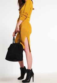 Femme dans une robe en tricot jaune moutarde avec une fente latérale, tenant un grand sac à main noir, portant des bottines à talons noirs, se tenant de côté.