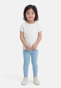 Mlad otrok s črnimi lasmi stoji obrnjeno naprej, oblečen v enobarvno belo majico, svetlo modre rebraste pajkice in bele superge.