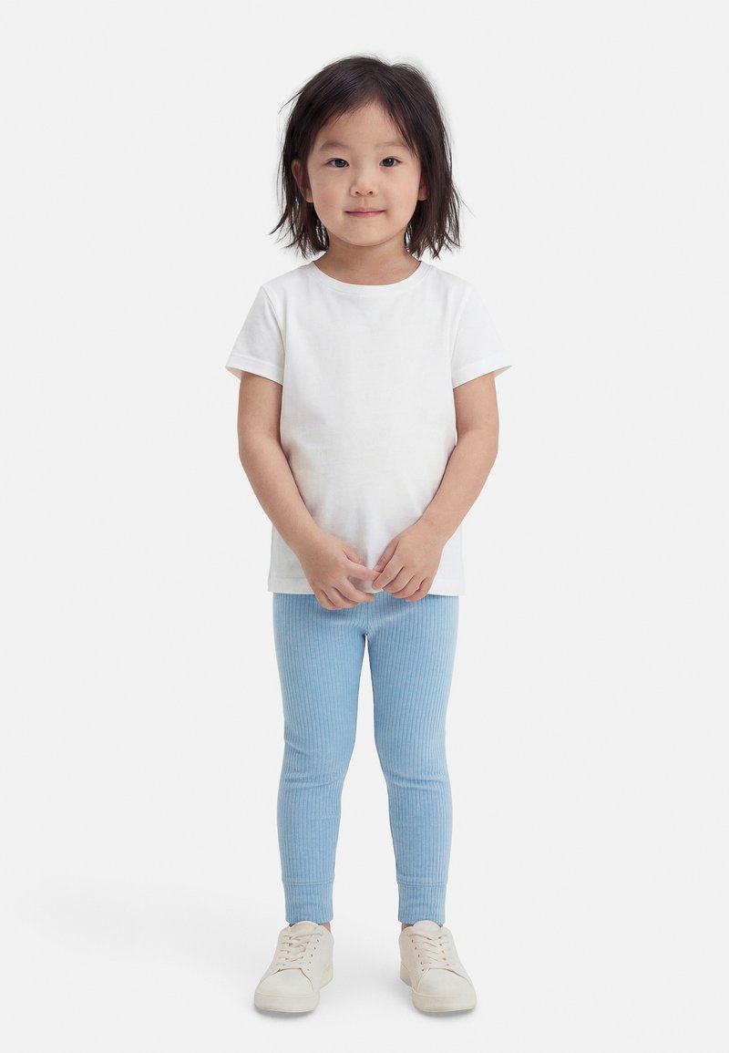 Mlad otrok s črnimi lasmi stoji obrnjeno naprej, oblečen v enobarvno belo majico, svetlo modre rebraste pajkice in bele superge.