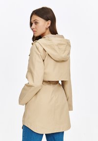 Beżowa krótka kurtka z kapturem, z paskiem w talii i szerokimi rękawami. Gładki materiał, minimalistyczny design i detal wentylacyjny z tyłu.
