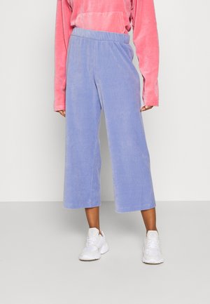 Person trägt hellblaue weite, kurze Hosen, ein rosafarbenes Langarmshirt und weiße Sportschuhe und steht auf einem grauen Boden.