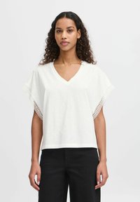 White V-neck t-shirt with short sleeves featuring intricate lace accents on edges. Soft, lightweight fabric; relaxed fit. Paired with black pants.