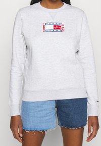 Sudadera gris claro con cuello redondo, con un logo bordado en rojo y azul. Combinada con shorts de mezclilla desgastados.