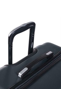 Maleta negra con ruedas, exterior texturizado, mango retráctil, asa lateral y cierre de cremallera. Presenta una etiqueta de marca en el mango.