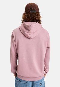 Sudadera con capucha rosa de corte relajado y puños acanalados, combinada con pantalones cortos de denim azul y una gorra marrón. Textura suave, diseño casual.