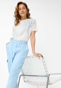 Light beige t-shirt with blue abstract pattern, paired with light blue relaxed pants. Model leans on a white woven chair with chrome accents.