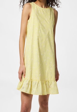 Femme portant une robe jaune sans manches avec broderie florale ajourée et un ourlet à volants, debout devant un fond uni.