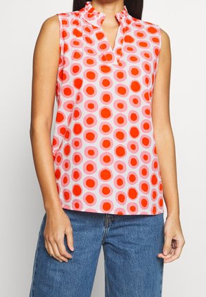 Top rosa senza maniche con motivi circolari rossi e rosa più chiaro, scollo arricciato, indossato con jeans blu.