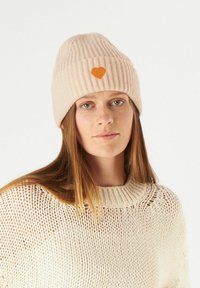 Lichtroze geribbelde gebreide beanie met een oranje hartvormige patch aan de voorkant. G gedragen met een beige gebreid trui met een losse structuur.
