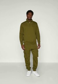 Vyras stovi su alyvuogių žalios spalvos užtrauktuku megztiniu, dėvėdamas cargo kelnes ir baltus sportbačius, prieš vienodą šviesią fono.