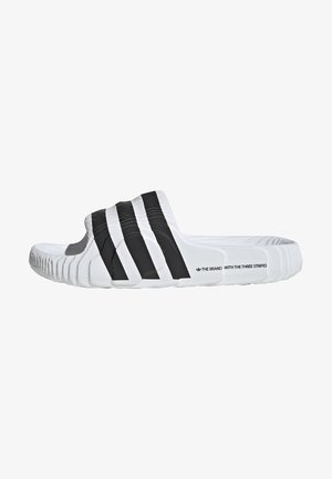 Klapki Adidas w białym i czarnym kolorze, z trzema czarnymi paskami na górze, teksturowaną podeszwą wewnętrzną i zakrzywioną podeszwą.
