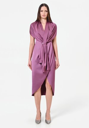 Femme portant une robe en satin violette sans manches avec un devant drapé noué par un détail en perle, associée à des talons pointus argentés.