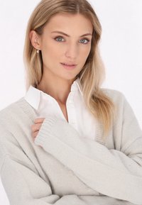 Světle šedý pletený kardigan s žebrovanými manžetami, přetetý přes bílou košili s límečkem, s jednoduchou texturou a uvolněným střihem.
