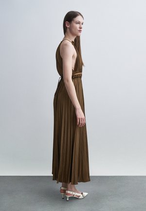 Femme debout de profil portant une robe marron plissée sans manches et des chaussures blanches à talons bas, sur un fond clair uni.