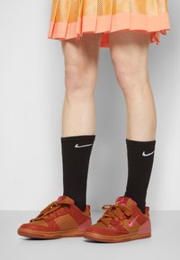 Baskets Nike présentant un mélange de matériaux rouge-orangé et marron avec une texture, agrémentées de chaussettes noires avec un logo blanc.