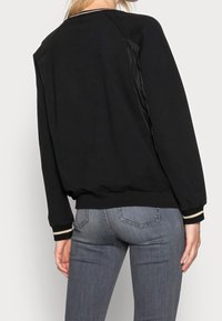 Zwarte sweatshirt met een relaxte pasvorm, voorzien van ribgebreide manchetten en een zoom, en een glanzend paneel op de schouders. Draagt over een paar grijze jeans.