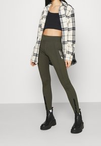 Olive drab legginsy z wysokim stanem i bocznymi detalami, zestawione z czarnym crop topem i koszulą w kratę, dopełnione czarnymi botkami.