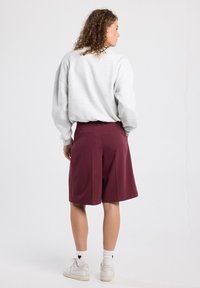 Shorts bordeaux au design ajusté, avec des plis à l'avant et des poches à l'arrière, portés avec un sweat-shirt oversize gris clair.