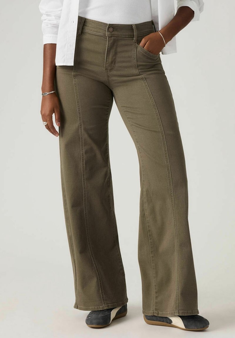 Personne portant un pantalon large vert olive, un haut blanc et des baskets foncées avec des rayures blanches, debout avec une main dans la poche.