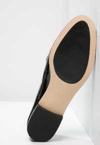 Semelle d'une chaussure habillée en cuir verni noir reposant contre un mur blanc, montrant des motifs de texture et le logo Michael Kors.