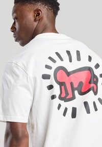 Valkoinen puuvillapaita, jossa on suuri grafiikka takana: punainen hahmo mustalla reunustettuna, jota ympäröivät säteilevät mustat viivat.