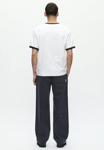 T-shirt bianco a maniche corte con colletto e bordo maniche verdi, abbinato a pantaloni larghi grigi scuri con tasche laterali.