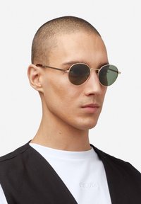 Gafas de sol redondas con lentes verdes, montura metálica fina y detalles de carey en las patillas. El modelo lleva una camiseta blanca debajo de un chaleco negro.