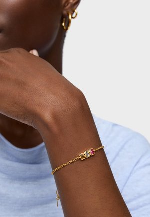 Złota bransoletka z łańcuszka z trzema kolorowymi zawieszkami — niebieską, różową i czarną — na tle o teksturowanym, brązowym odcieniu skóry.