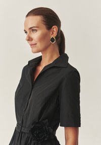 Čierne šaty s krátkymi rukávmi, golierom a zapínaním na gombíky. Obsahujú textúrovaný kvetinový akcent v oblasti pása. Hladká tkanina.