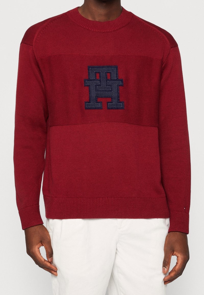 Homme portant un pull en maille rouge foncé avec un logo géométrique cousu en bleu marine sur la poitrine, assorti à un pantalon blanc.